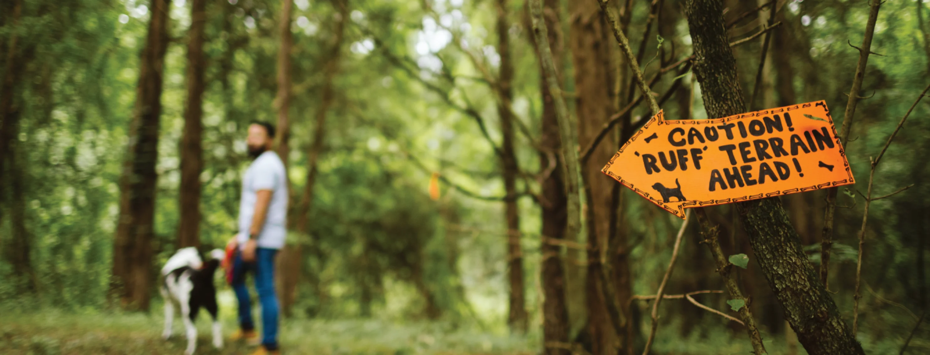 A man walking his black and white dog in the forest with a caution arrow sign that says, "Caution Ruff Terrain Ahead!" in bright orange. 